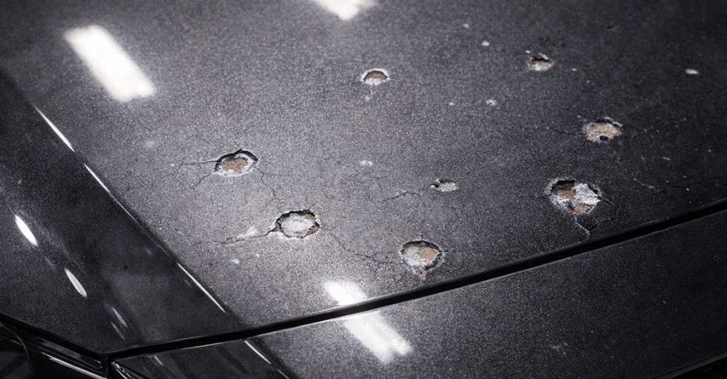 Close-up photorealistic view of a dark metallic car hood with multiple rock chips exposing primer and bare metal, surrounded by cracked and slightly oxidized paint, under natural outdoor lighting, illustrating paint damage that paint protection film helps prevent.